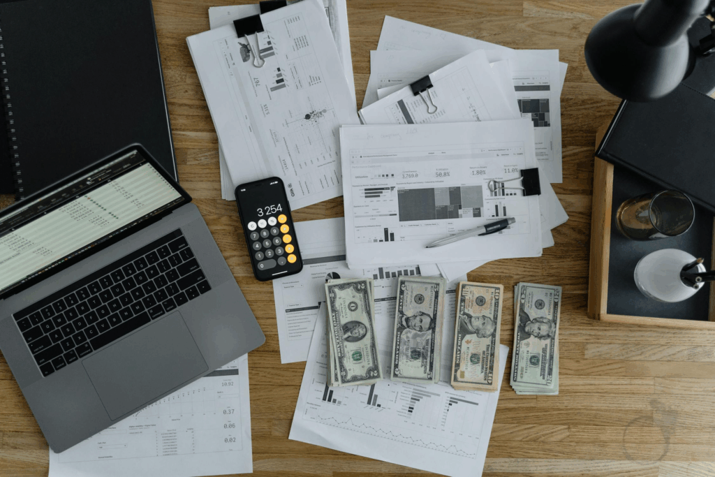 The image depicts a cluttered desk filled with financial documents, colorful charts, and printed tables. A laptop screen displays a detailed spreadsheet, while a smartphone calculator shows the number 3,254. Several stacks of U.S. dollar bills lie nearby, along with a sleek black notebook, a pen, a desk lamp, and a glass of water — all elements that suggest an intense session of financial planning or budgeting. This visual evokes the complexity and preparation often involved in securing a mortgage in Spain, especially for foreign buyers navigating unfamiliar financial systems.