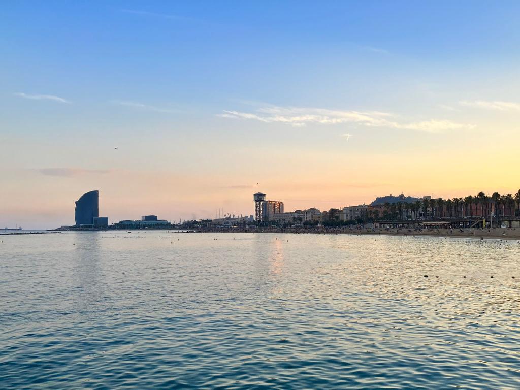 This photo captures a serene sunset over the Barcelona coastline. You can see the iconic W Barcelona Hotel (sail-shaped building) on the left, a calm sea in the foreground, and the cityscape with palm trees and beachgoers stretching along the shore to the right. The sky transitions from warm hues near the horizon to soft blue above, reflecting beautifully on the water.