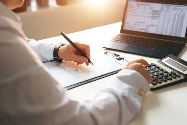 This image shows a person engaged in financial or administrative work. They are sitting at a desk with documents, a pen in one hand, and using a calculator with the other. A laptop displaying spreadsheets or financial data is also open in front of them. The setting suggests a professional environment, likely related to accounting, bookkeeping, budgeting, or financial analysis. The lighting and overall tone imply a focused and diligent working atmosphere.
