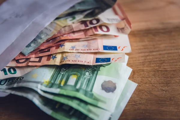 The image shows several euro banknotes partially inside a white envelope, placed on a wooden surface. The visible denominations include €10, €20, and €100 notes. The money appears to be fanned out, suggesting it has either just been placed into or taken out of the envelope. This kind of image often represents cash savings, a financial transaction, or money being given or received discreetly.