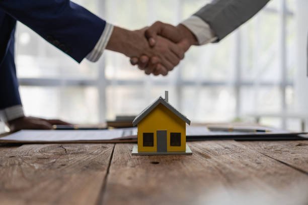 his image illustrates a real estate transaction or agreement. It features two individuals shaking hands across a table, symbolizing a successful deal or partnership. In the foreground, there's a small model of a yellow house, indicating that the agreement is related to buying, selling, or investing in property. The presence of documents on the table further suggests a formal contract or negotiation process.