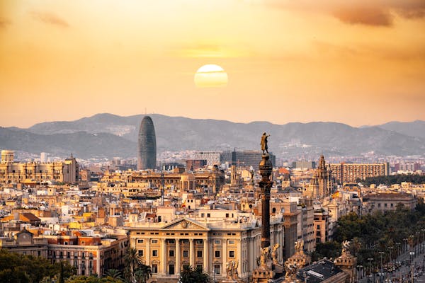 This striking photo captures a sunset view over the city of Barcelona, Spain. The iconic Columbus Monument (pointing out to sea) stands prominently in the foreground, while the distinctive Torre Glòries (formerly Torre Agbar) rises in the background. The sun sets over the distant Collserola hills, casting a warm golden glow over the densely packed rooftops and historic architecture of the city.