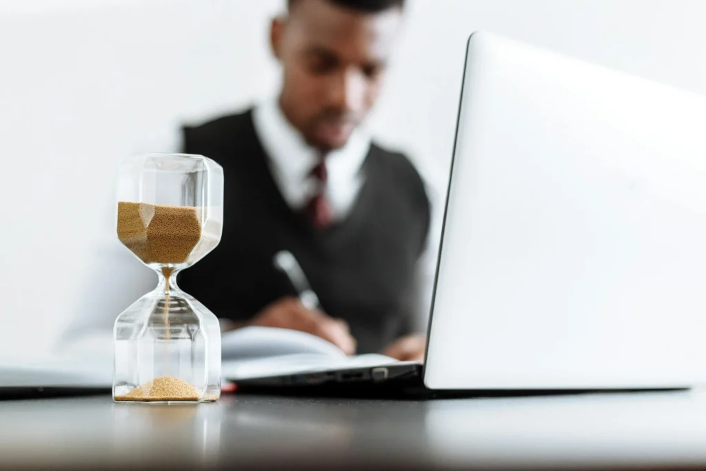 This image conveys the concept of time management or productivity. It shows a clear hourglass in the foreground, with sand visibly passing through, symbolizing the passage of time. In the background, a professional man is working at a desk with a laptop and notebook, slightly out of focus, suggesting concentration and task execution. The hourglass serves as a metaphor for deadlines, efficient work habits, or the value of time in a professional setting.